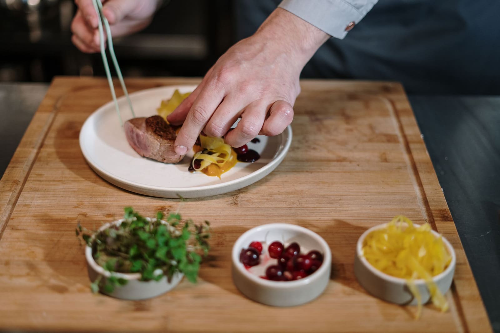 A chef plating a dish on a wooden counter, prepared garnishes lined up in bowls beside it.