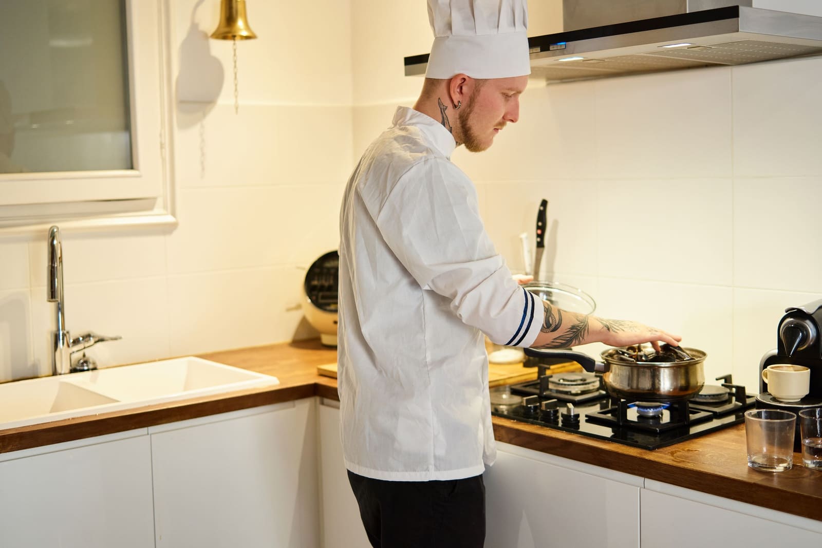 A chef in whites cooking at the hob of a bright contemporary kitchen, daylight from the window behind.
