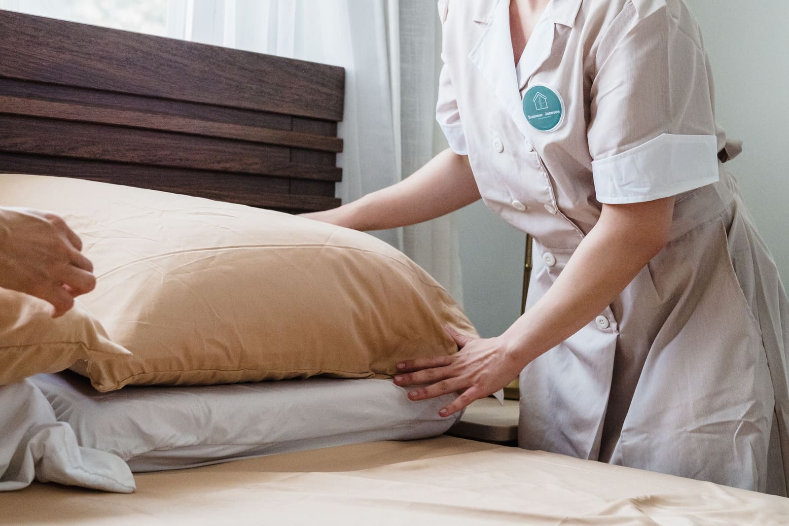 A housekeeper in uniform plumping pillows on a freshly made bed in a bright room.