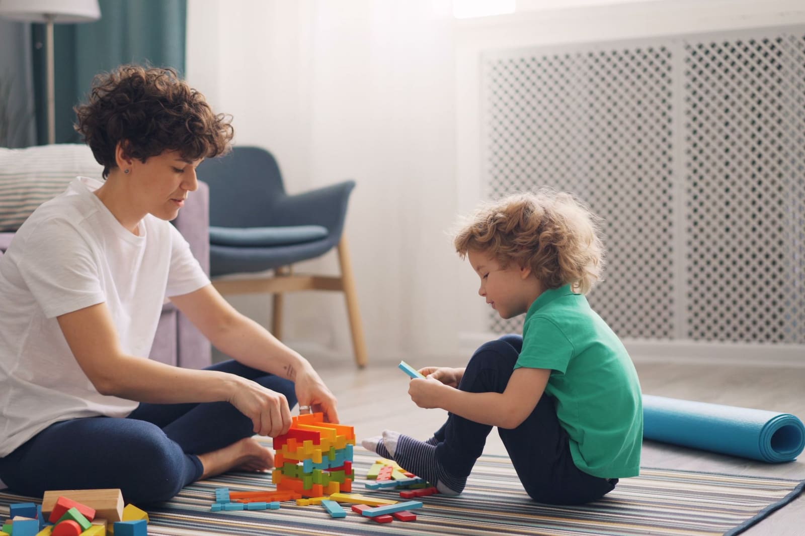 A nanny and a young child building together with blocks on the floor of a light, calm living room.