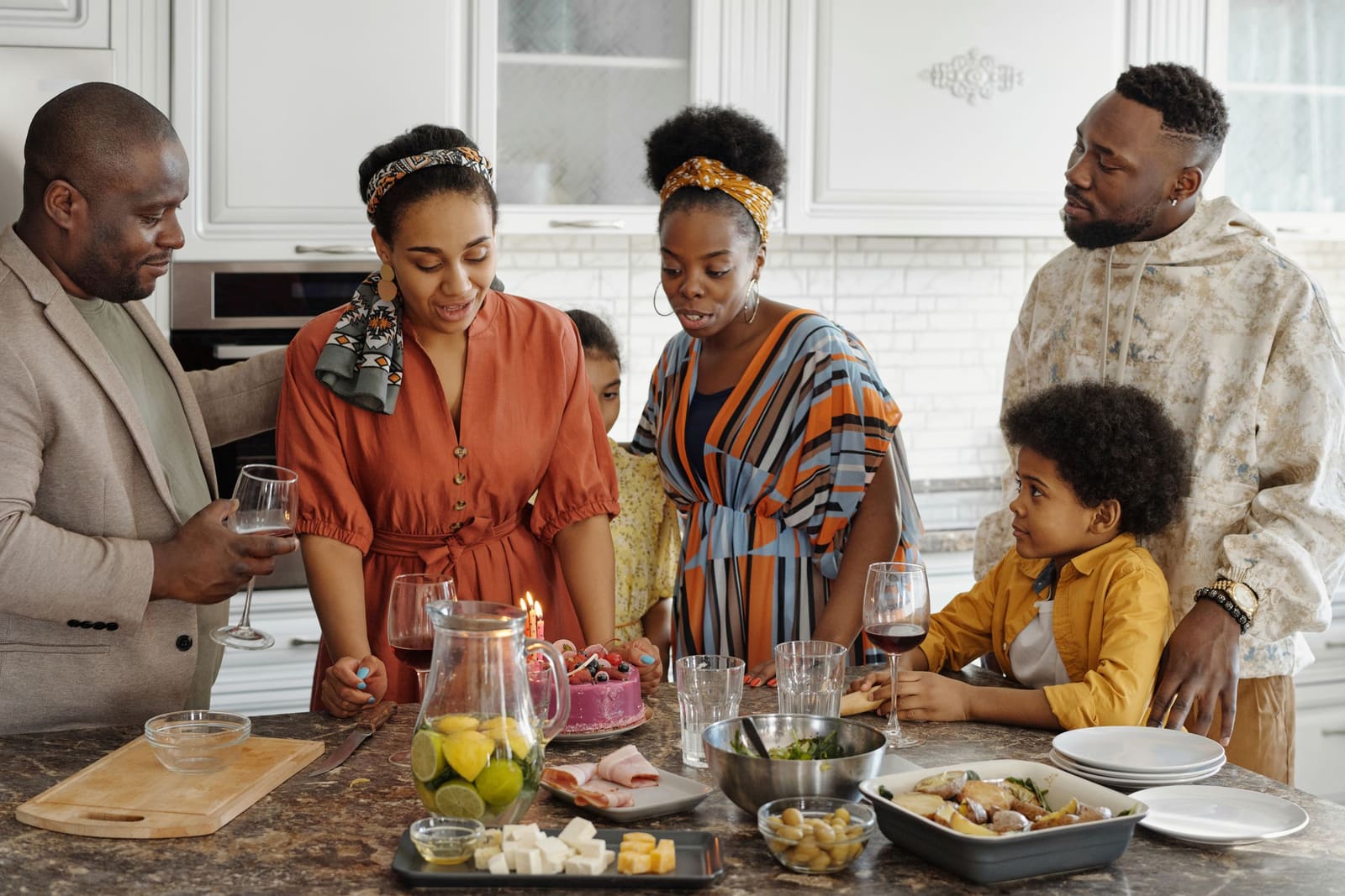 A family and their guests gathered round the island of a bright modern kitchen, food and glasses laid out between them.