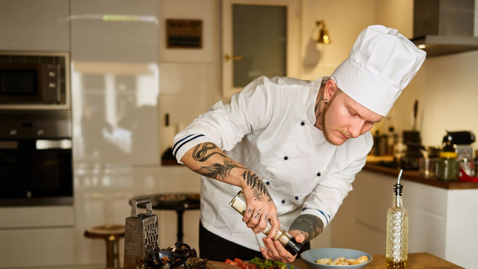 A private chef in whites seasoning a dish he is plating on the counter of a bright contemporary home kitchen.