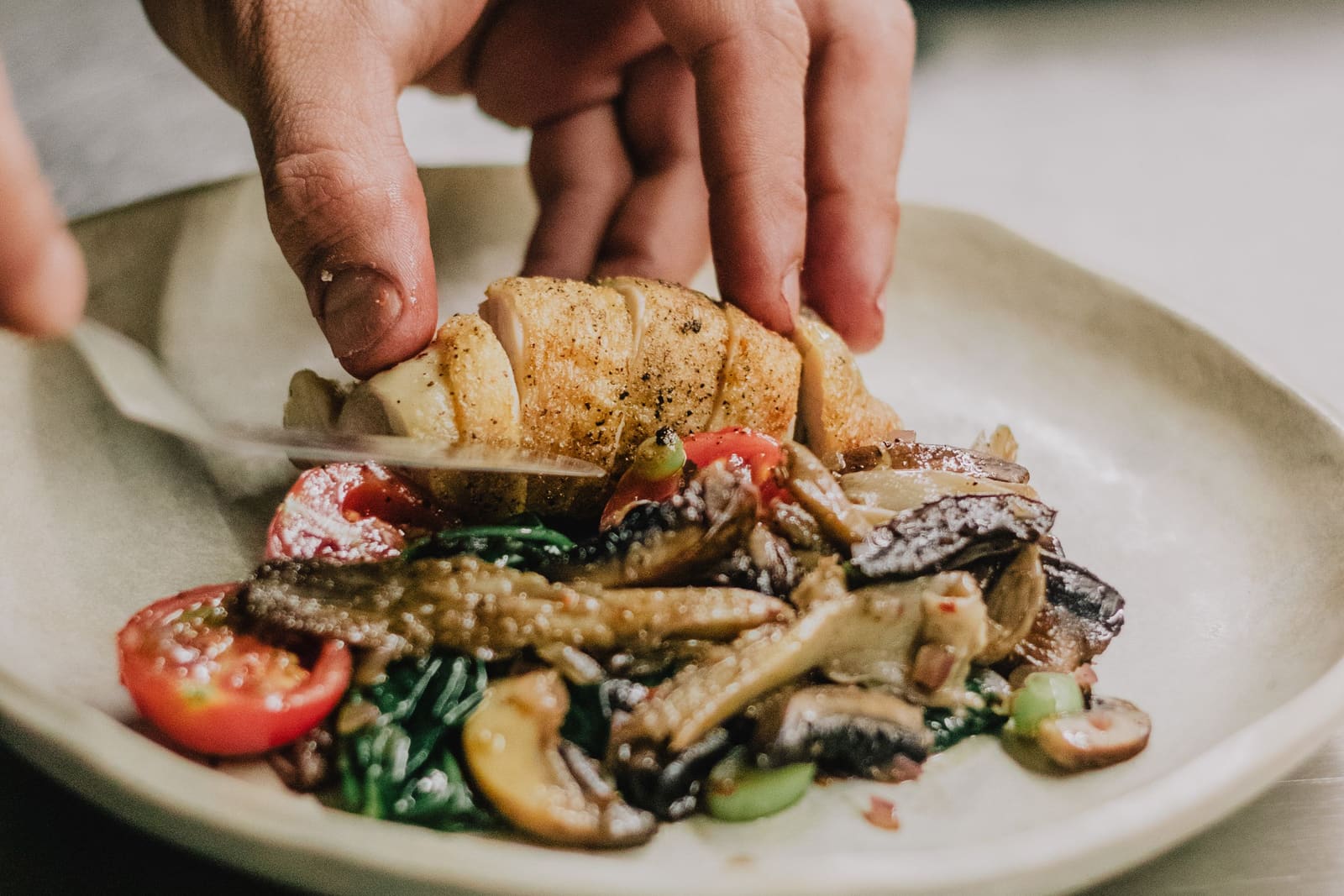A chef's hands laying sliced chicken over roasted vegetables, tomatoes and greens on a plate.